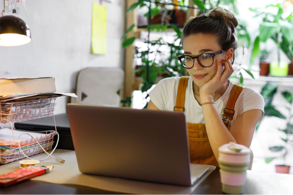 Young woman in overalls on work from home arrangements, wearing glasses looking at a laptop computer. She has a cup of coffee.